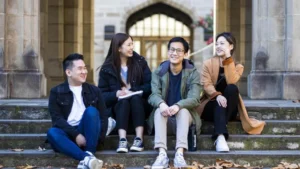 Four young adults sitting on outdoor steps, laughing and chatting, in front of a university building