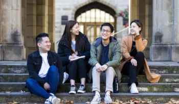 Four young adults sitting on outdoor steps, laughing and chatting, in front of a university building