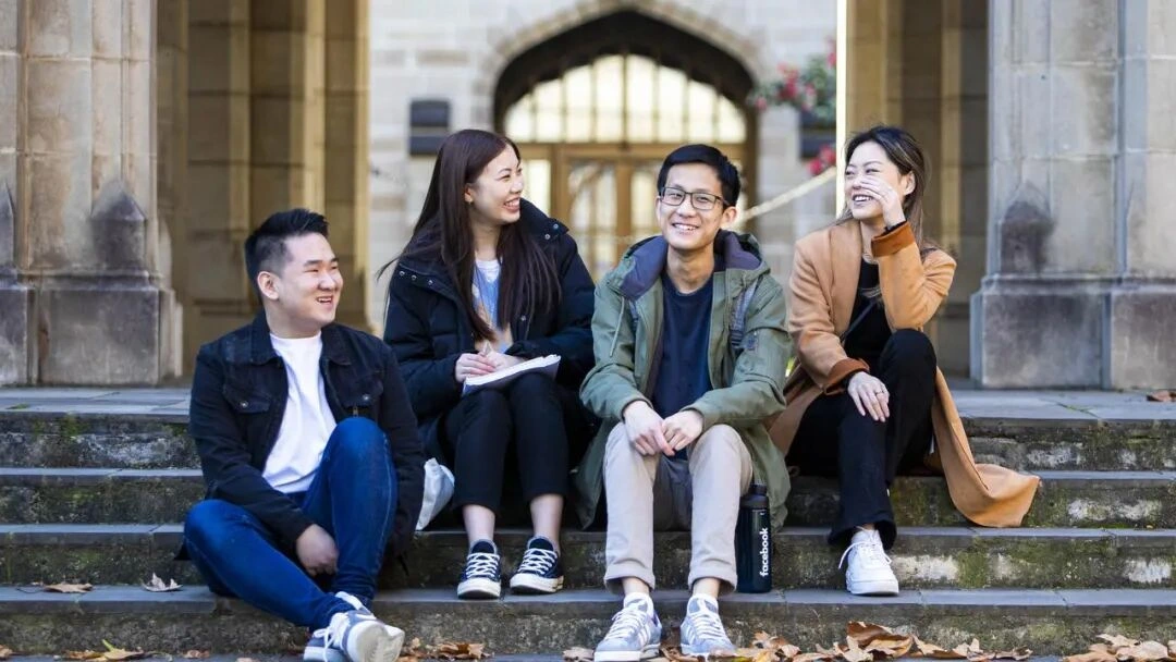 Four young adults sitting on outdoor steps, laughing and chatting, in front of a university building
